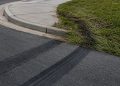 Close-up of fresh tire marks curving toward a sidewalk curb beside flattened grass.