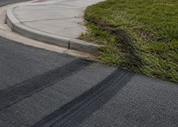 Close-up of fresh tire marks curving toward a sidewalk curb beside flattened grass.