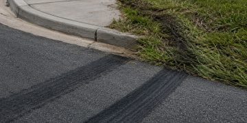 Close-up of fresh tire marks curving toward a sidewalk curb beside flattened grass.