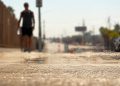 Close-up of heat waves shimmering above sun-baked pavement, with a blurred pedestrian carrying water beneath intense afternoon sunlight.