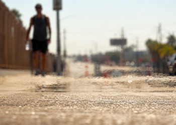 Close-up of heat waves shimmering above sun-baked pavement, with a blurred pedestrian carrying water beneath intense afternoon sunlight.