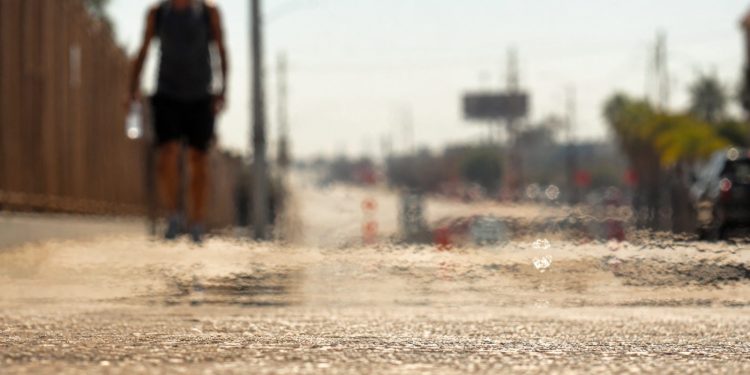 Close-up of heat waves shimmering above sun-baked pavement, with a blurred pedestrian carrying water beneath intense afternoon sunlight.