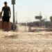 Close-up of heat waves shimmering above sun-baked pavement, with a blurred pedestrian carrying water beneath intense afternoon sunlight.