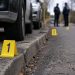 Close-up of police evidence markers near a curb and parked vehicles, with two blurred investigators standing farther down the street.