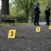 Close-up of police evidence markers near a park walkway with blurred officers standing in the background.