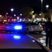 Close-up of police lights reflecting across a grocery store parking lot at night, with a blurred officer standing near parked cars.