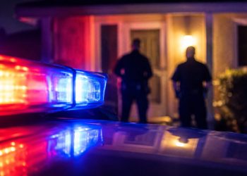 Close-up of red and blue emergency light reflections outside a residential doorway at night, with two blurred officers standing farther back.
