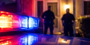 Close-up of red and blue emergency light reflections outside a residential doorway at night, with two blurred officers standing farther back.