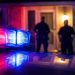 Close-up of red and blue emergency light reflections outside a residential doorway at night, with two blurred officers standing farther back.