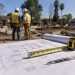 Close-up of residential rebuilding plans and measuring tools on a cleared lot, with two blurred construction workers standing in the background.