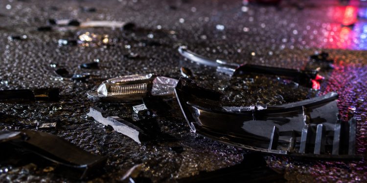Close-up of shattered vehicle debris and broken plastic pieces scattered across dark pavement at night.