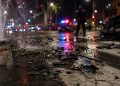 Close-up of shattered vehicle debris and water flowing across an intersection at night, with a blurred officer and emergency lights in the background.