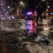 Close-up of shattered vehicle debris and water flowing across an intersection at night, with a blurred officer and emergency lights in the background.