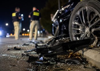 Close-up of shattered vehicle debris beside a damaged wheel at night, with two blurred investigators standing near the roadway in the background.