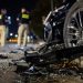 Close-up of shattered vehicle debris beside a damaged wheel at night, with two blurred investigators standing near the roadway in the background.