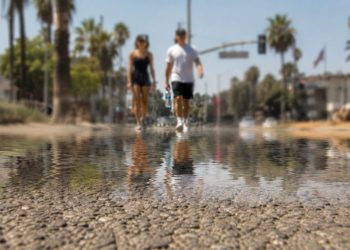 Close-up of shimmering heat above sun-baked pavement, with two blurred pedestrians carrying water in the distant background.