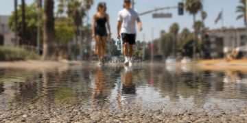 Close-up of shimmering heat above sun-baked pavement, with two blurred pedestrians carrying water in the distant background.