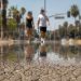 Close-up of shimmering heat above sun-baked pavement, with two blurred pedestrians carrying water in the distant background.