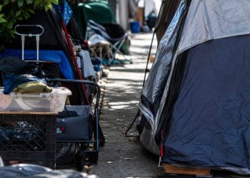 Close-up of tents and personal belongings narrowing a residential sidewalk, with no identifiable homes or street signs.