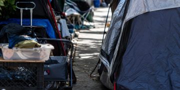 Close-up of tents and personal belongings narrowing a residential sidewalk, with no identifiable homes or street signs.