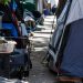 Close-up of tents and personal belongings narrowing a residential sidewalk, with no identifiable homes or street signs.