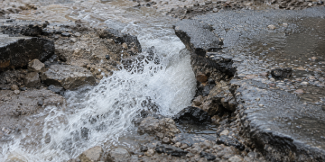 Close-up of water forcefully rushing through broken asphalt and muddy debris, used as an illustrative image for a Hollywood Hills water main break that damaged part of a roadway.