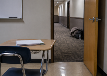 Empty classroom desk with an open door leading to a blurred hotel-style hallway, where personal belongings rest along the corridor wall.