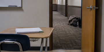 Empty classroom desk with an open door leading to a blurred hotel-style hallway, where personal belongings rest along the corridor wall.