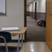 Empty classroom desk with an open door leading to a blurred hotel-style hallway, where personal belongings rest along the corridor wall.