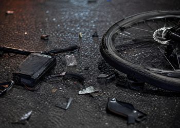 lose-up of shattered bicycle parts scattered across dark asphalt beside a damaged wheel.