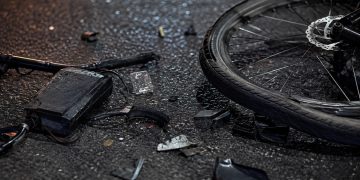 lose-up of shattered bicycle parts scattered across dark asphalt beside a damaged wheel.