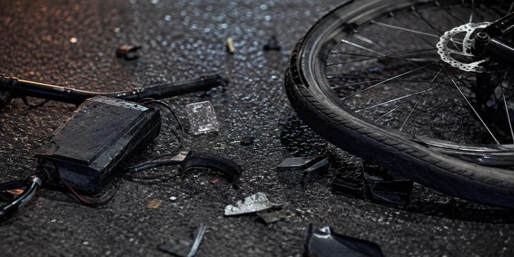 lose-up of shattered bicycle parts scattered across dark asphalt beside a damaged wheel.