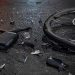 lose-up of shattered bicycle parts scattered across dark asphalt beside a damaged wheel.