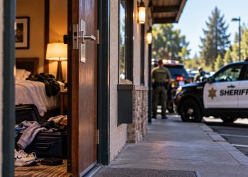 Big Bear hotel room investigation with an open hotel doorway, scattered personal belongings and blurred sheriff's vehicles outside.