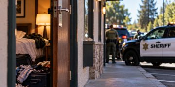 Big Bear hotel room investigation with an open hotel doorway, scattered personal belongings and blurred sheriff's vehicles outside.