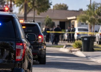 Blurred police vehicles and officers outside a San Bernardino home during an officer-involved shooting investigation, with the residential street in the background.