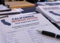 Close-up of California ballot initiative petitions and signature forms on a table, with blurred paperwork and a pen illustrating an alleged election fraud investigation.