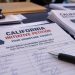 Close-up of California ballot initiative petitions and signature forms on a table, with blurred paperwork and a pen illustrating an alleged election fraud investigation.
