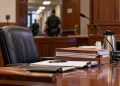 Close-up of a California courtroom defense table with legal files and an empty attorney chair, with a courthouse corridor blurred in the background.
