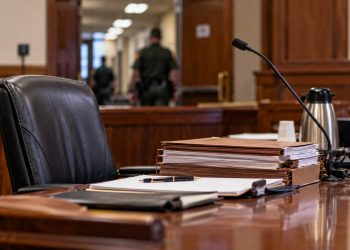 Close-up of a California courtroom defense table with legal files and an empty attorney chair, with a courthouse corridor blurred in the background.