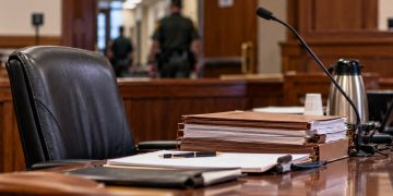 Close-up of a California courtroom defense table with legal files and an empty attorney chair, with a courthouse corridor blurred in the background.