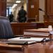 Close-up of a California courtroom defense table with legal files and an empty attorney chair, with a courthouse corridor blurred in the background.