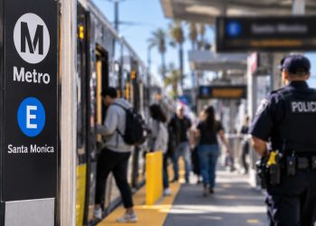 Close-up of a Metro E Line platform with blurred passengers and a uniformed police officer in the background, illustrating expanded transit patrols in Santa Monica.