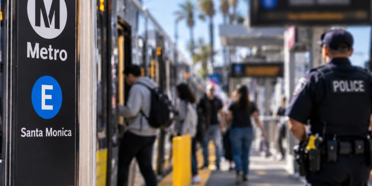 Close-up of a Metro E Line platform with blurred passengers and a uniformed police officer in the background, illustrating expanded transit patrols in Santa Monica.