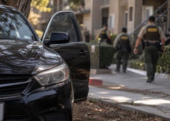 Close-up of a black sedan abandoned near an apartment complex, with blurred sheriff's deputies searching the area in the background.
