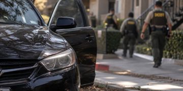 Close-up of a black sedan abandoned near an apartment complex, with blurred sheriff's deputies searching the area in the background.