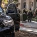 Close-up of a black sedan abandoned near an apartment complex, with blurred sheriff's deputies searching the area in the background.