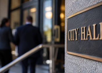 Close-up of a blurred municipal building entrance with indistinct figures in the background, illustrating an investigation involving a city official.