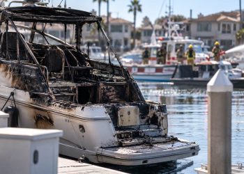 Close-up of a charred boat in a Huntington Harbour residential slip, with firefighters and harbor vessels blurred in the background.