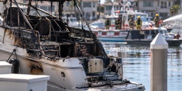 Close-up of a charred boat in a Huntington Harbour residential slip, with firefighters and harbor vessels blurred in the background.